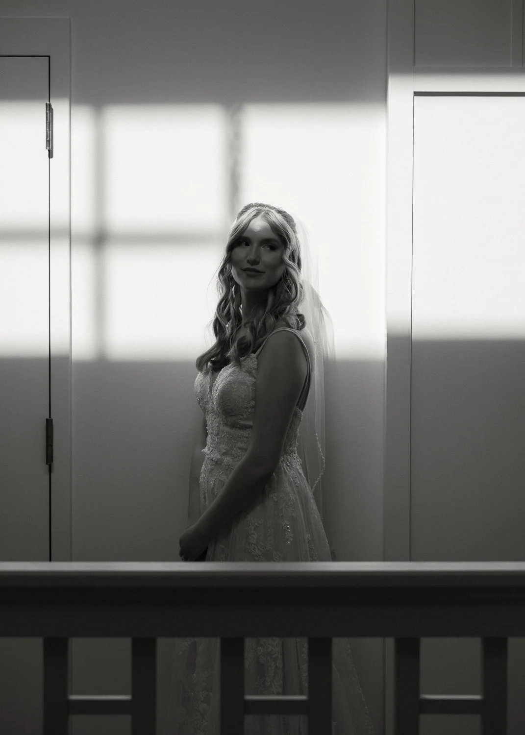 A woman in a wedding dress, standing indoors with sunlight creating shadow patterns on the wall behind her.