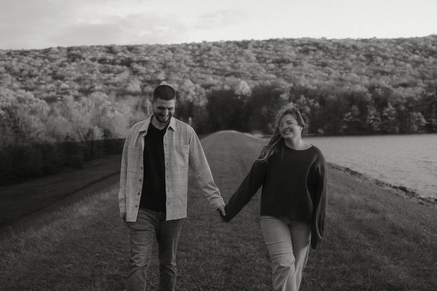A black and white photo of a smiling couple walking hand in hand outdoors near a lake and wooded hills.
