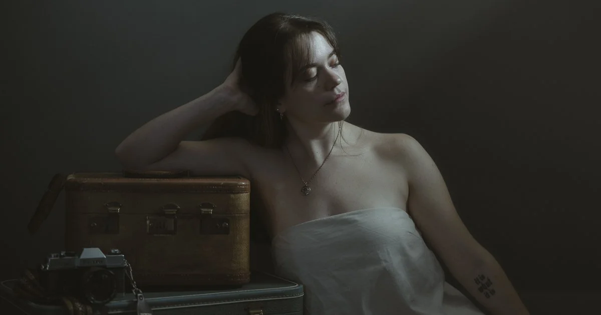 A woman with auburn hair and a tattoo on her arm, sitting with her eyes closed and resting her hand behind her head, appears to be wrapped in a white towel or sheet, with a vintage suitcase, camera, and another suitcase in front of her, against a dark background.