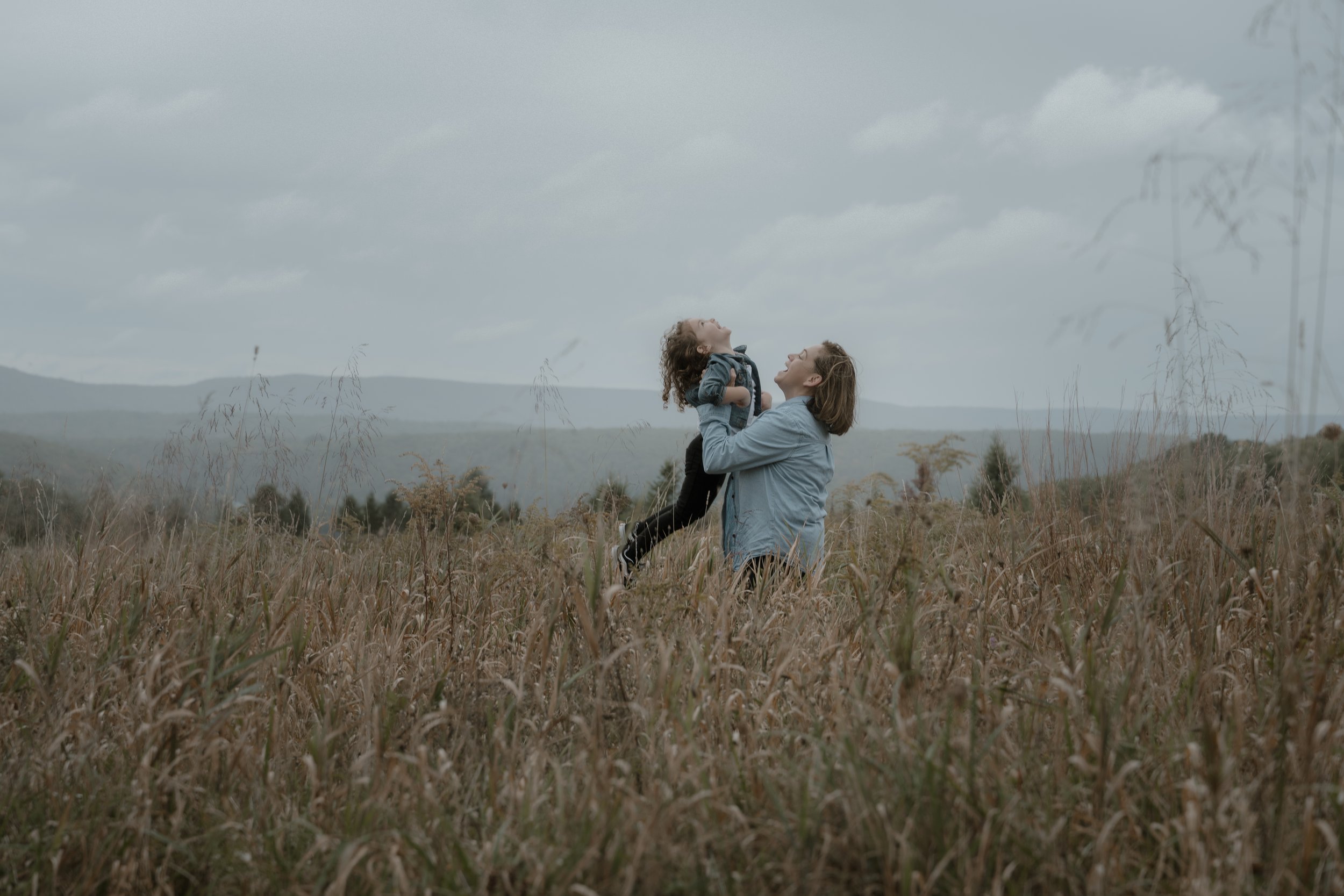 A woman lifting a young girl in a grassy field with mountains and cloudy sky in the background.