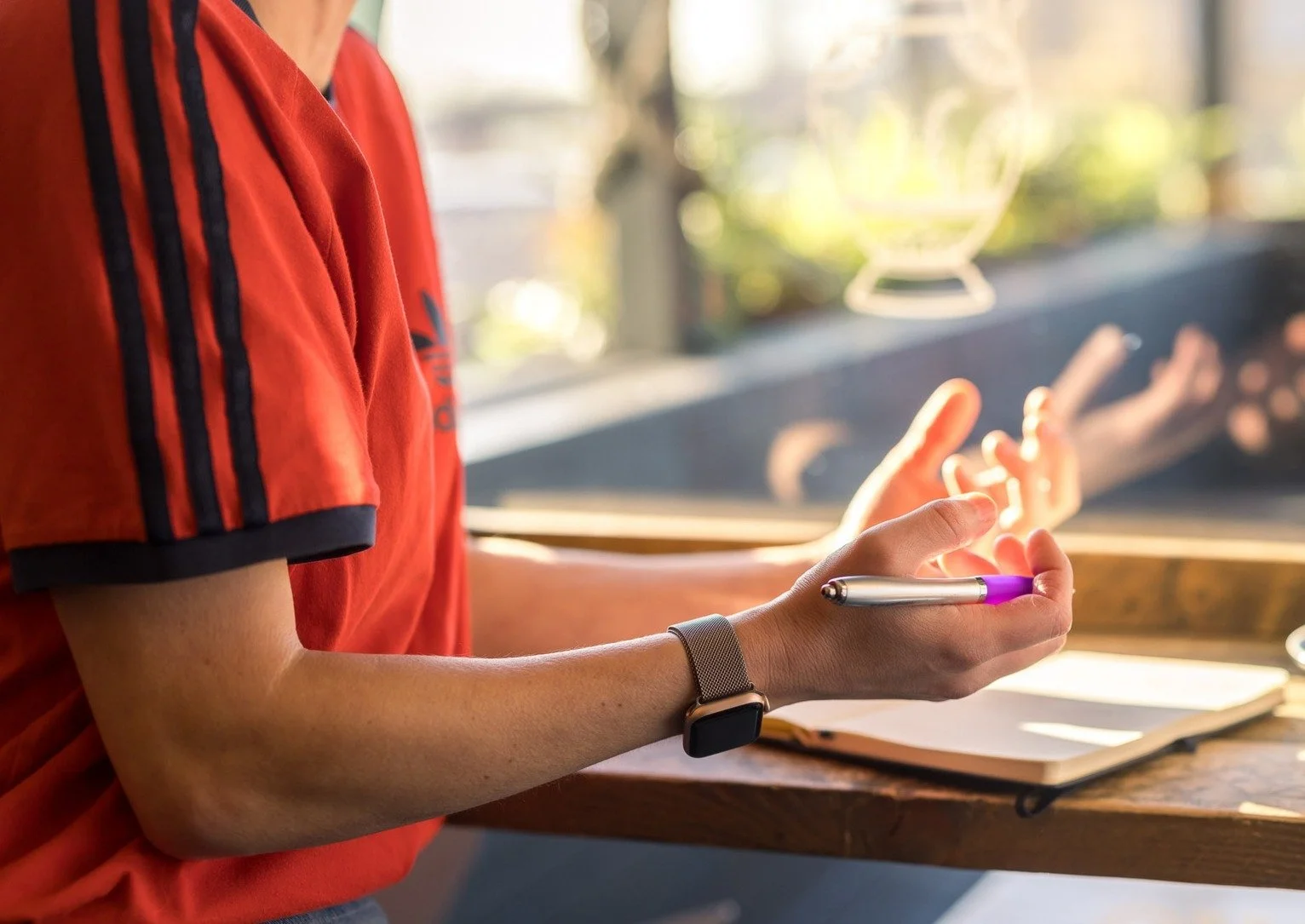 A person in a red Adidas shirt holding a purple and silver pen near an open notebook on a wooden surface, with sunshine and blurred plants in the background.