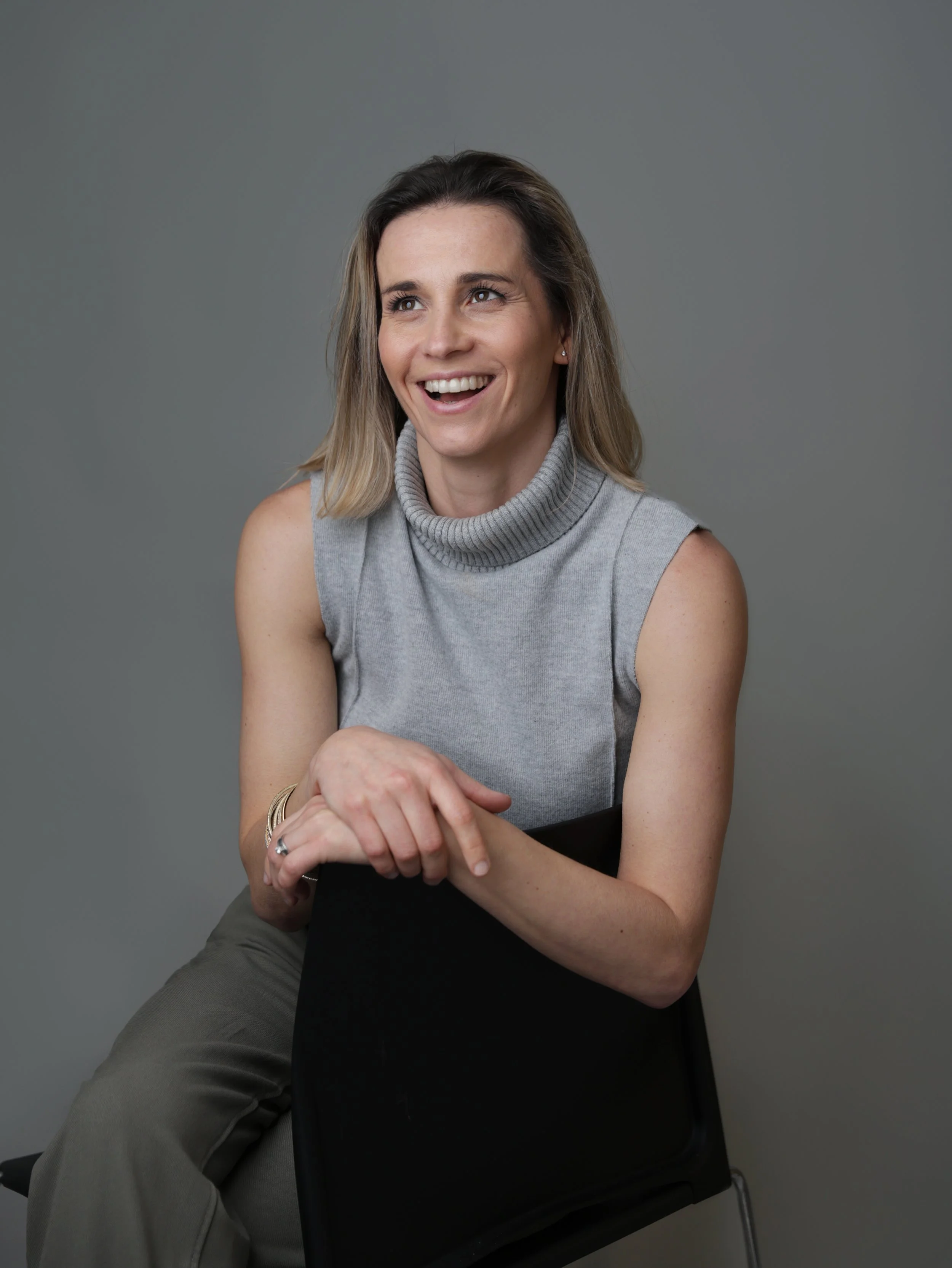A woman with long, light brown hair, smiling and looking to her right, sitting on a black chair against a plain gray background. She is wearing a sleeveless gray turtleneck top and khaki pants.