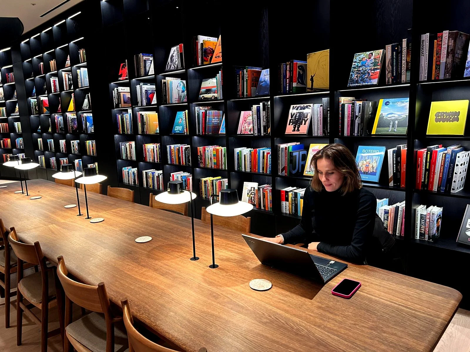 A woman sitting at a long wooden table in a bookstore or library, working on a laptop with bookshelves behind her.