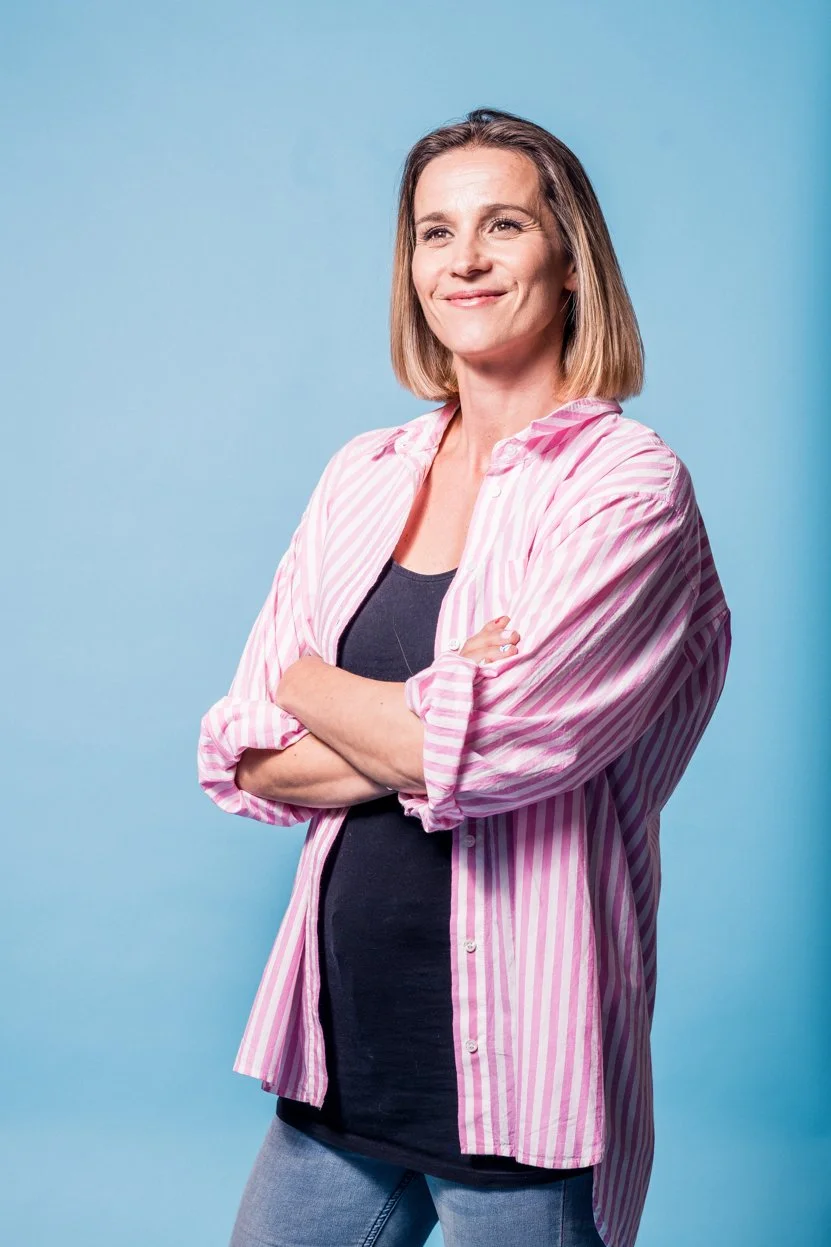 A woman with shoulder-length brown hair smiling confidently with arms crossed, wearing a pink and white striped shirt over a black top, standing against a light blue background.