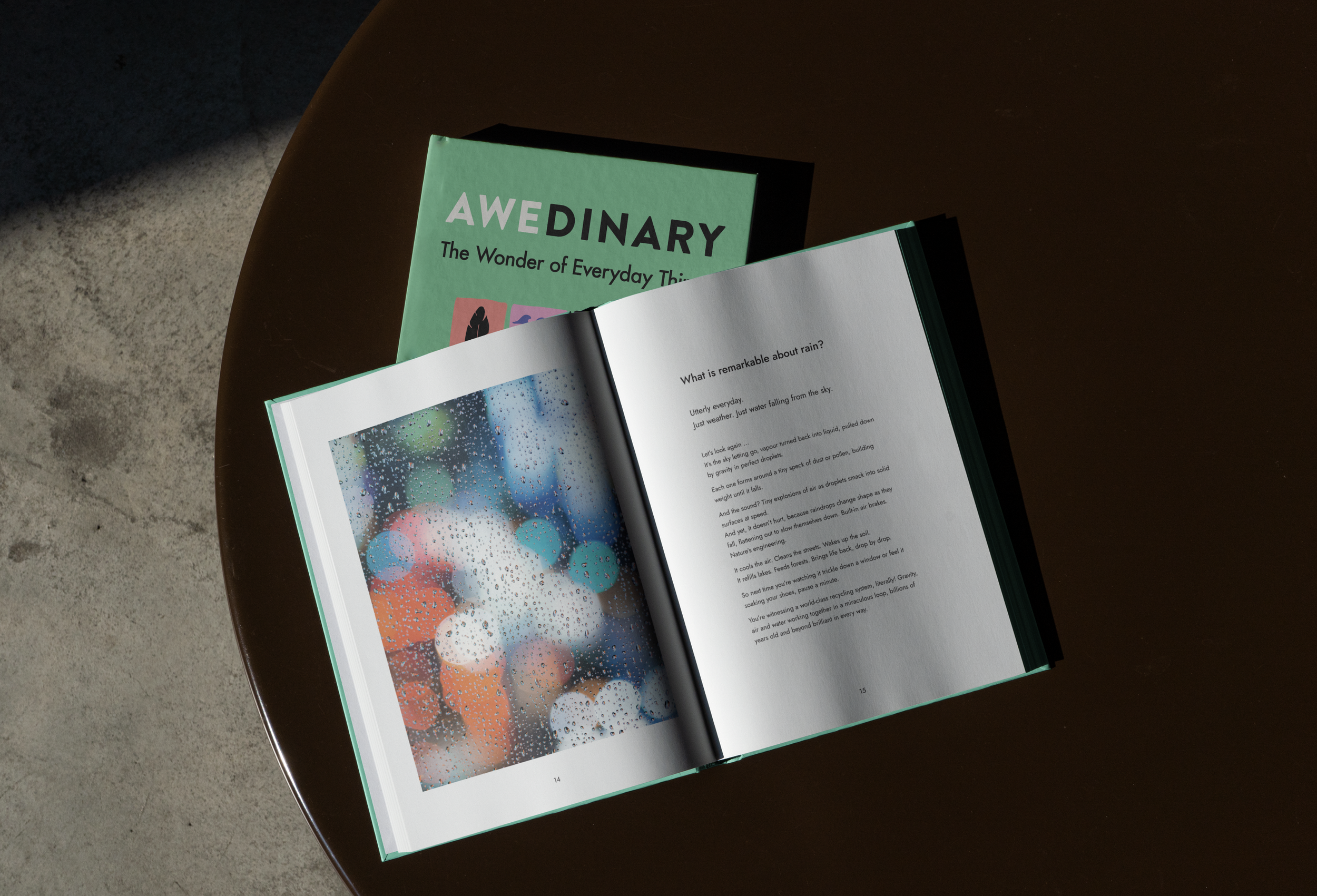 An open book with the title 'AWEDINARY The Wonder of Everyday Things' and a page showing a poem about rain, placed on a round wooden table. Behind it, a closed book with the same title.