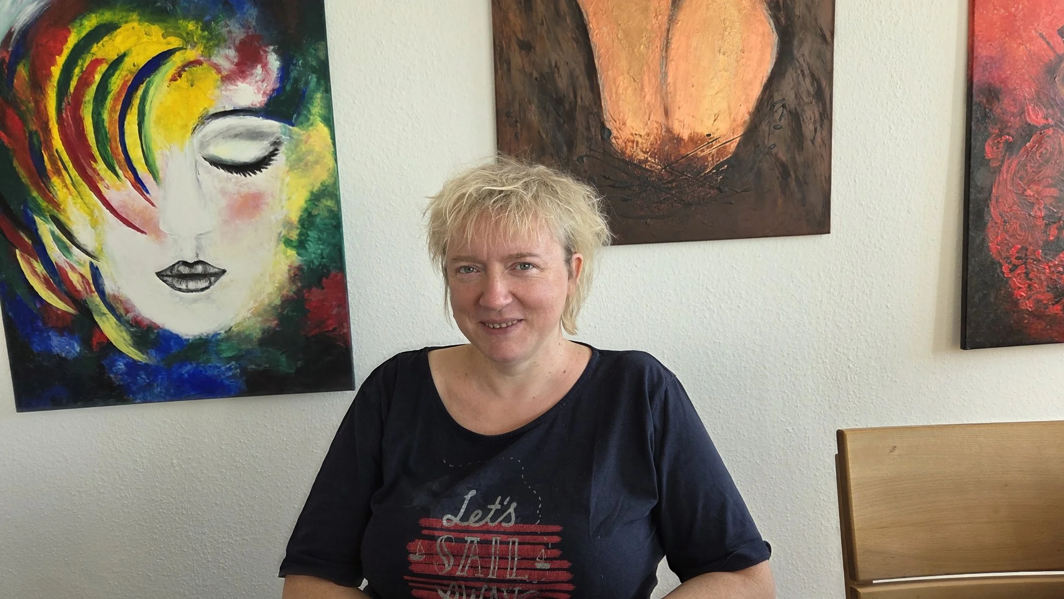 Eine Frau sitzt lachend in einem Raum vor mehreren bunten abstrakten Kunstwerken an der Wand.