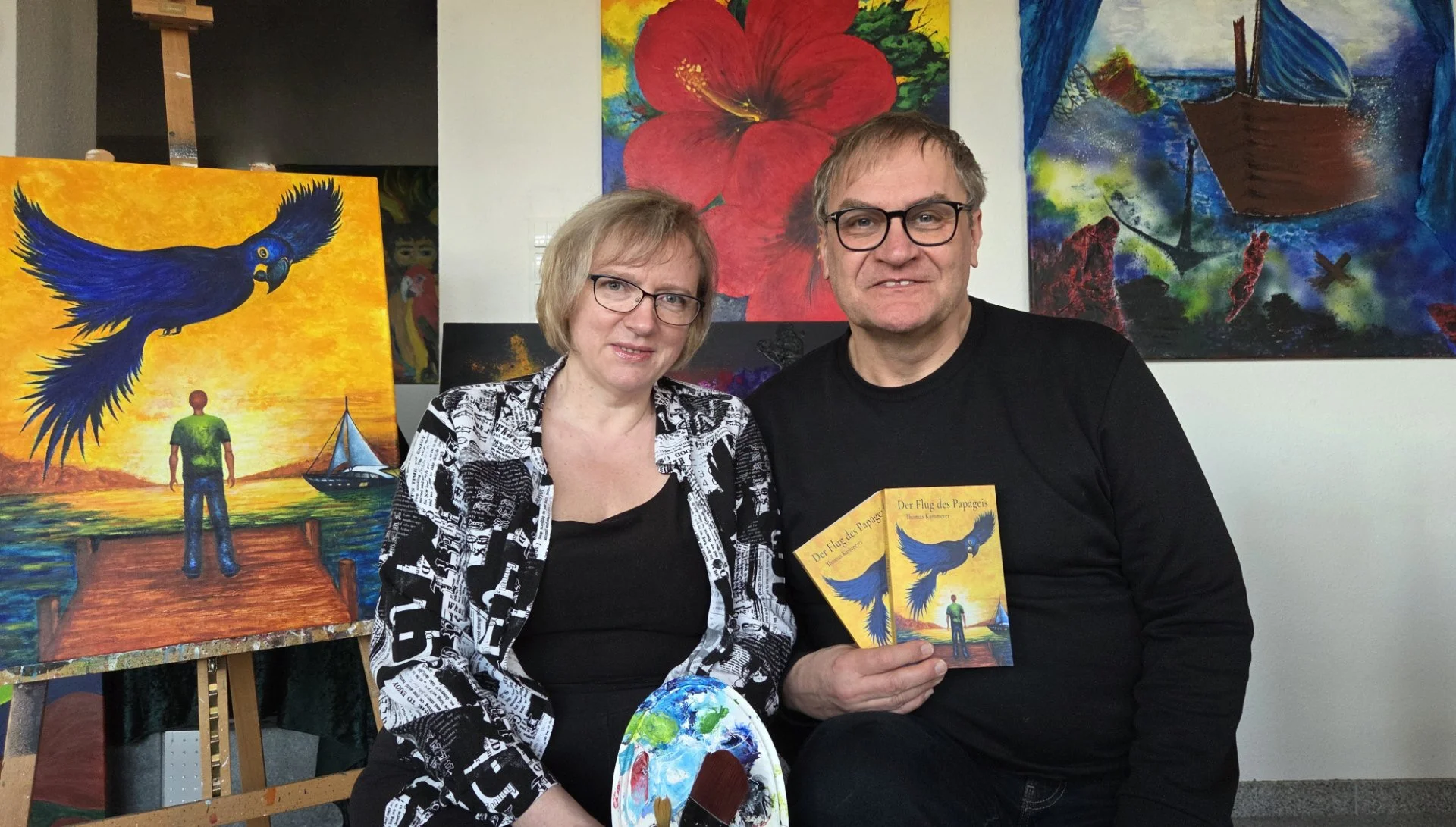 Ein Mann und eine Frau sitzen vor farbenfrohen Gemälden, die einen Himmel, ein Schiff, einen großen roten Hibiskus und einen fliegenden Papagei zeigen, während der Mann ein Buch mit Titel 'Der Flug des Papageis' hält.