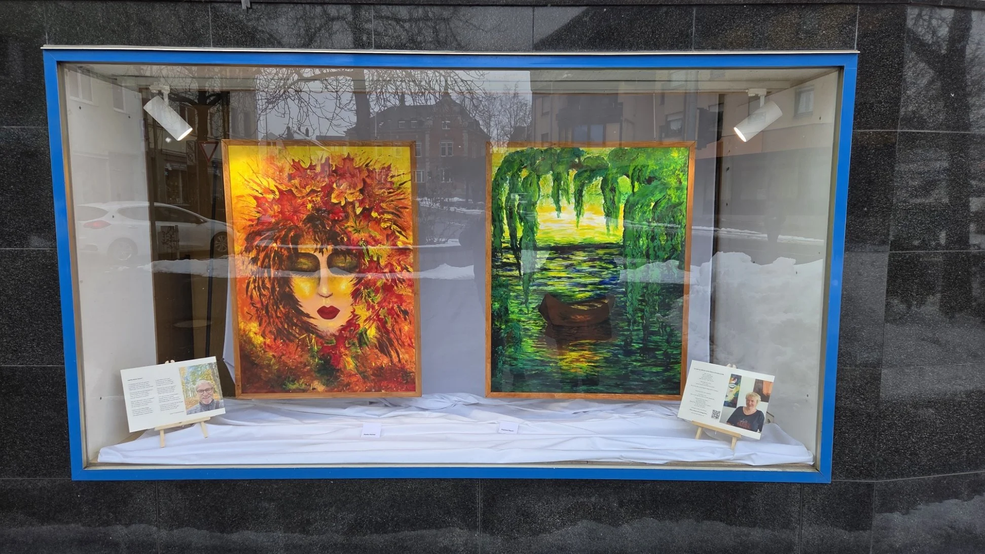 Schaufenster mit zwei Gemälden, ein Porträt mit roten und orangefarbenen Blättern im Haar und eine Szenerie mit einem Boot auf einem Fluss im Grünen, mit informationalen Schildern darunter.