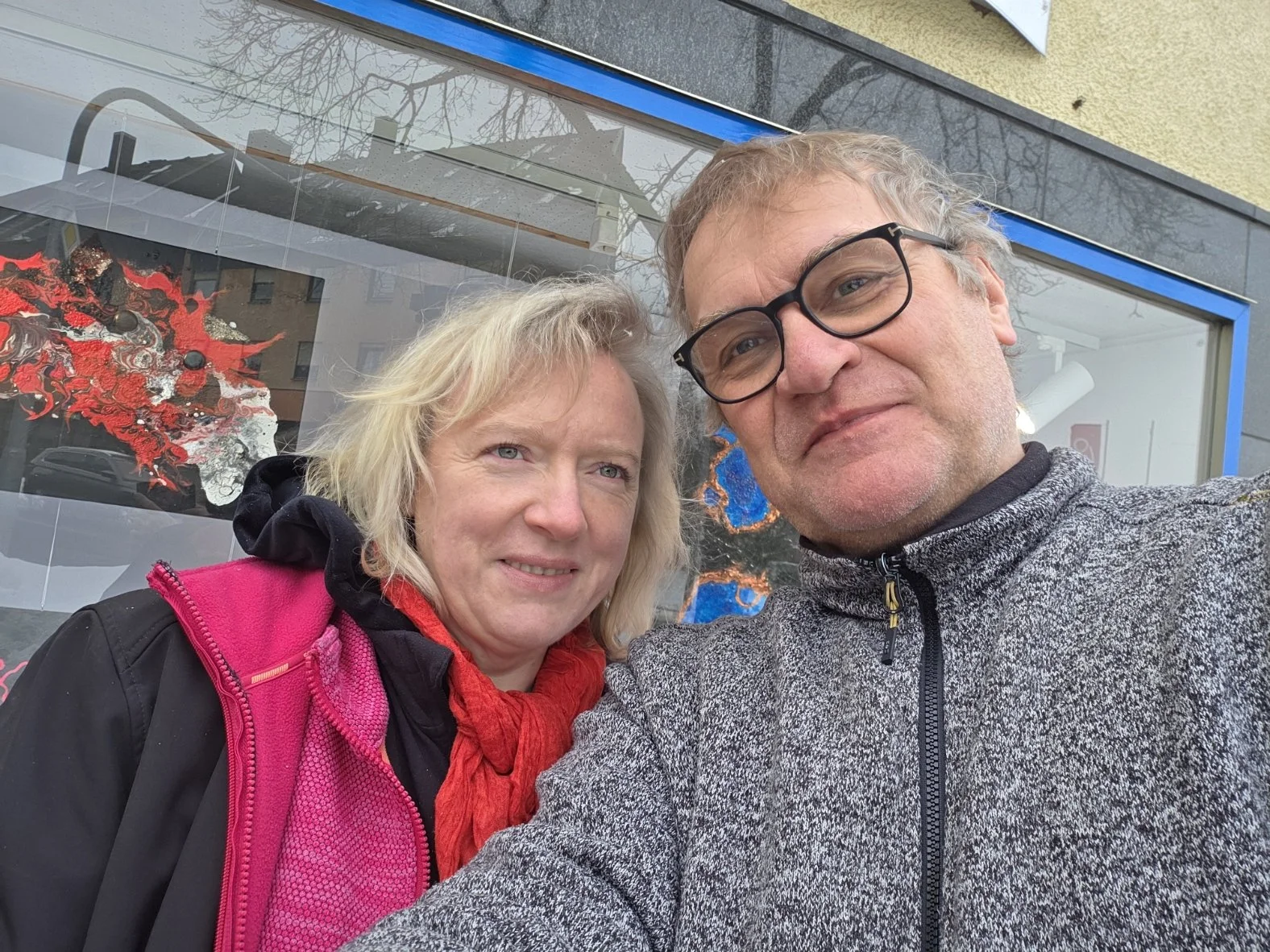 Ein Mann mit Brille und eine Frau fotografieren sich vor einem Kunstgeschäft. Im Hintergrund sind bunte Kunstwerke zu sehen, wahrscheinlich Gemälde oder Skulpturen.