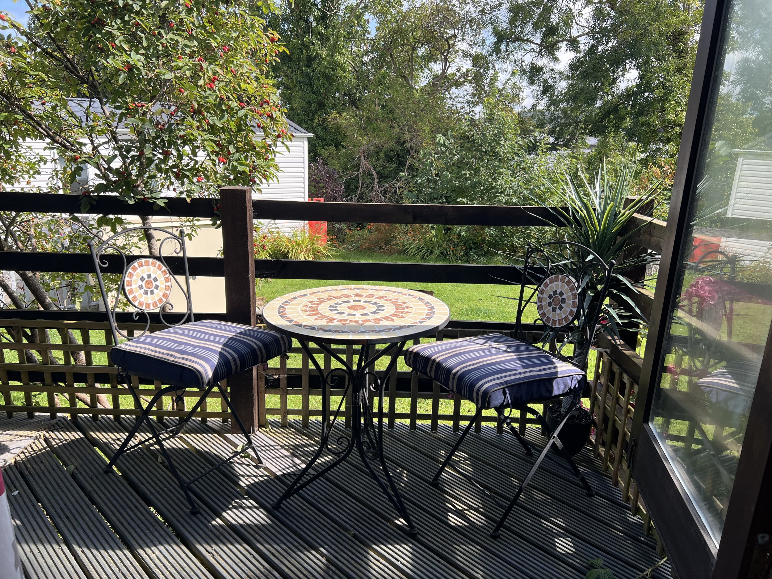 A small patio with a round mosaic table and two metal chairs with striped cushions, overlooking a green lawn and surrounded by trees and bushes.
