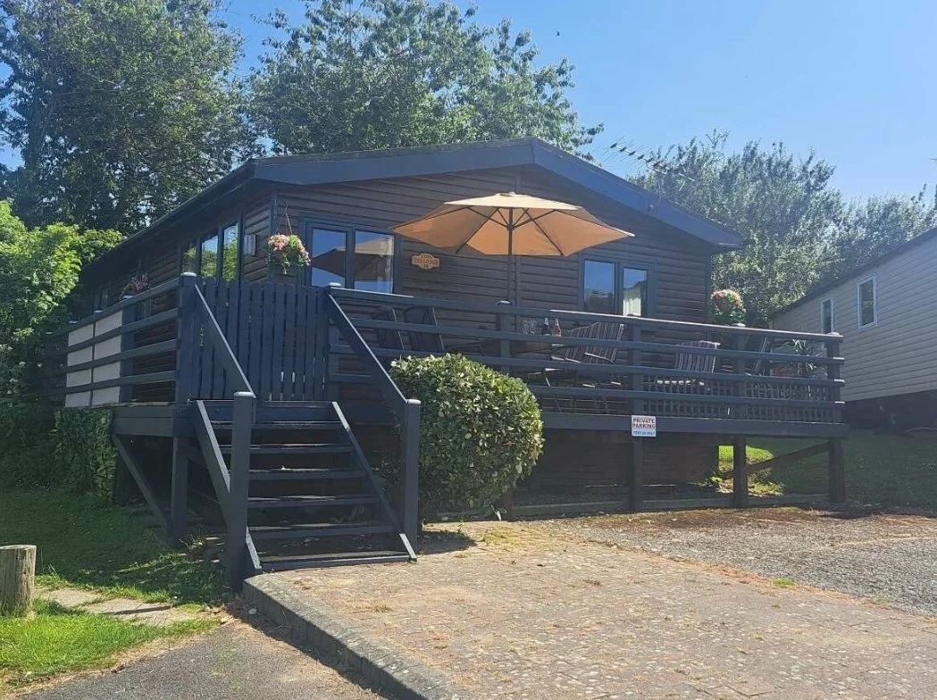 A dark wooden house with a deck, stairs, and an umbrella outside, surrounded by trees and neighbouring houses.