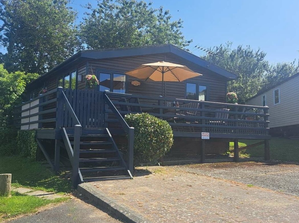 A small dark-colored house with a deck and stairs, outdoor furniture including a table, chairs, and an umbrella, and decorative flower pots on the railing, with trees and a neighboring house in the background.
