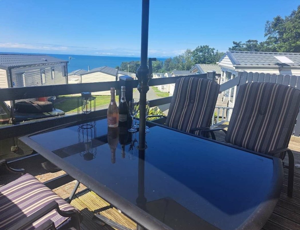Outdoor patio with a glass table, three bottles of wine or champagne, two glasses, striped outdoor chairs, and a view of houses, trees, and the ocean in the background on a sunny day.