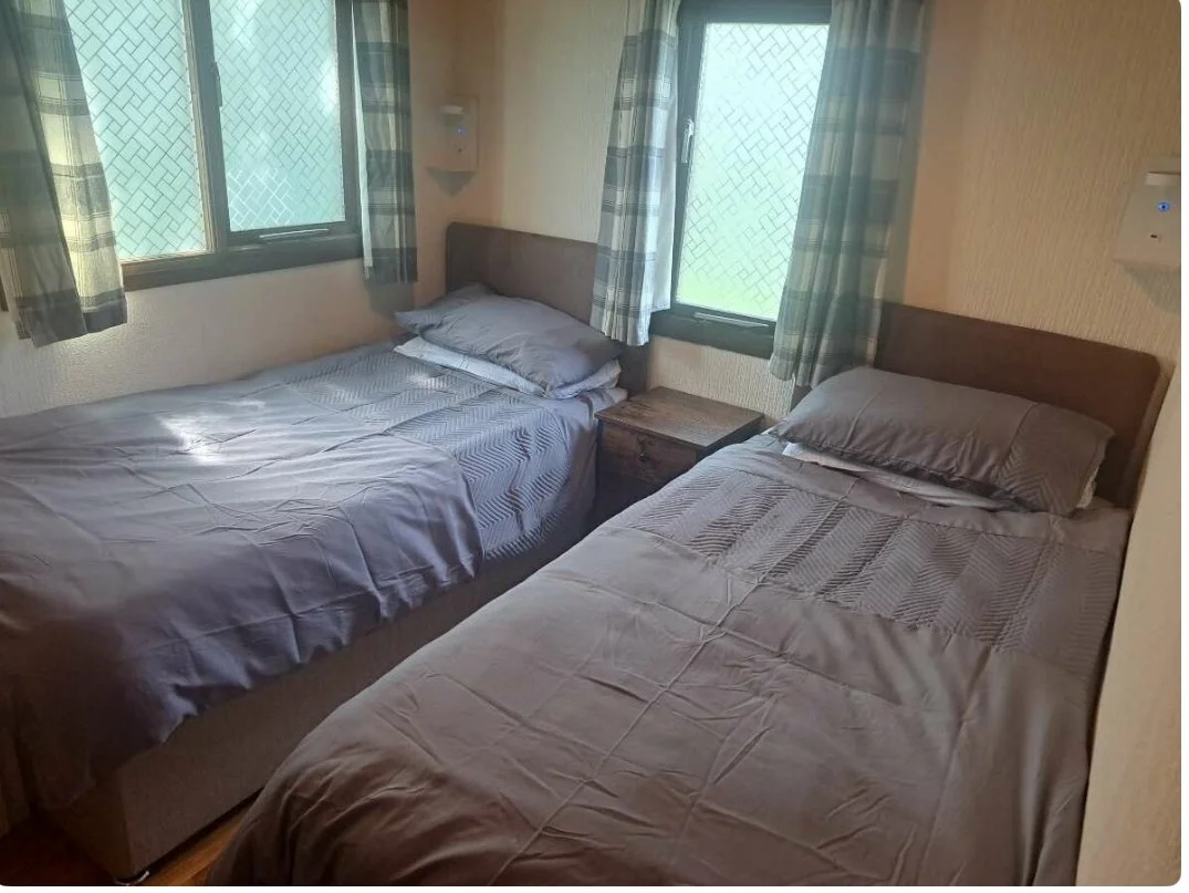 Twin beds with gray bedding and pillows, a nightstand between them, with windows covered by patterned curtains in a bedroom.