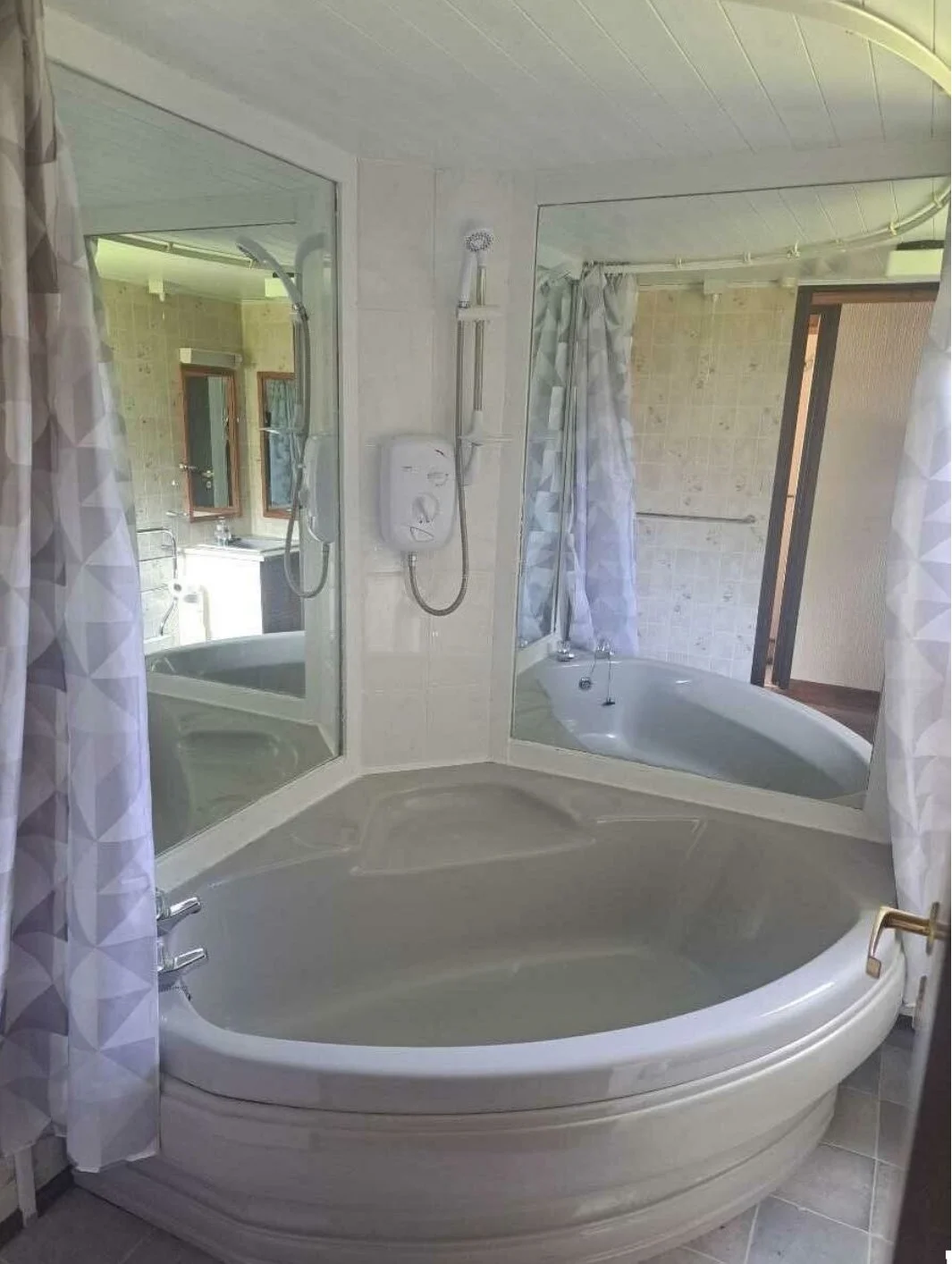 A bathroom with a corner bathtub, a glass wall with a shower head, a mirror reflecting the bathtub and wall, and shower curtains on the sides.