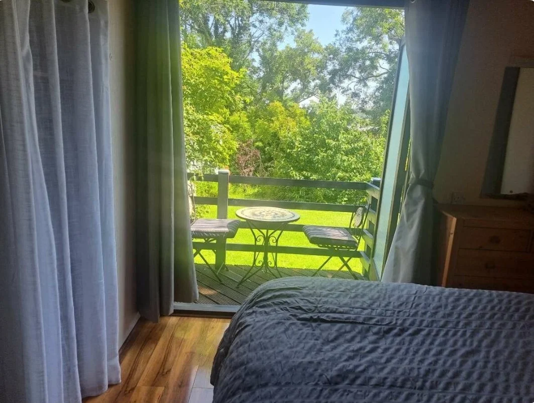 View of a balcony with a round metal table and two chairs, overlooking green trees and a grassy yard, seen from inside a bedroom with a part of a bed, curtains, and wooden furniture visible.