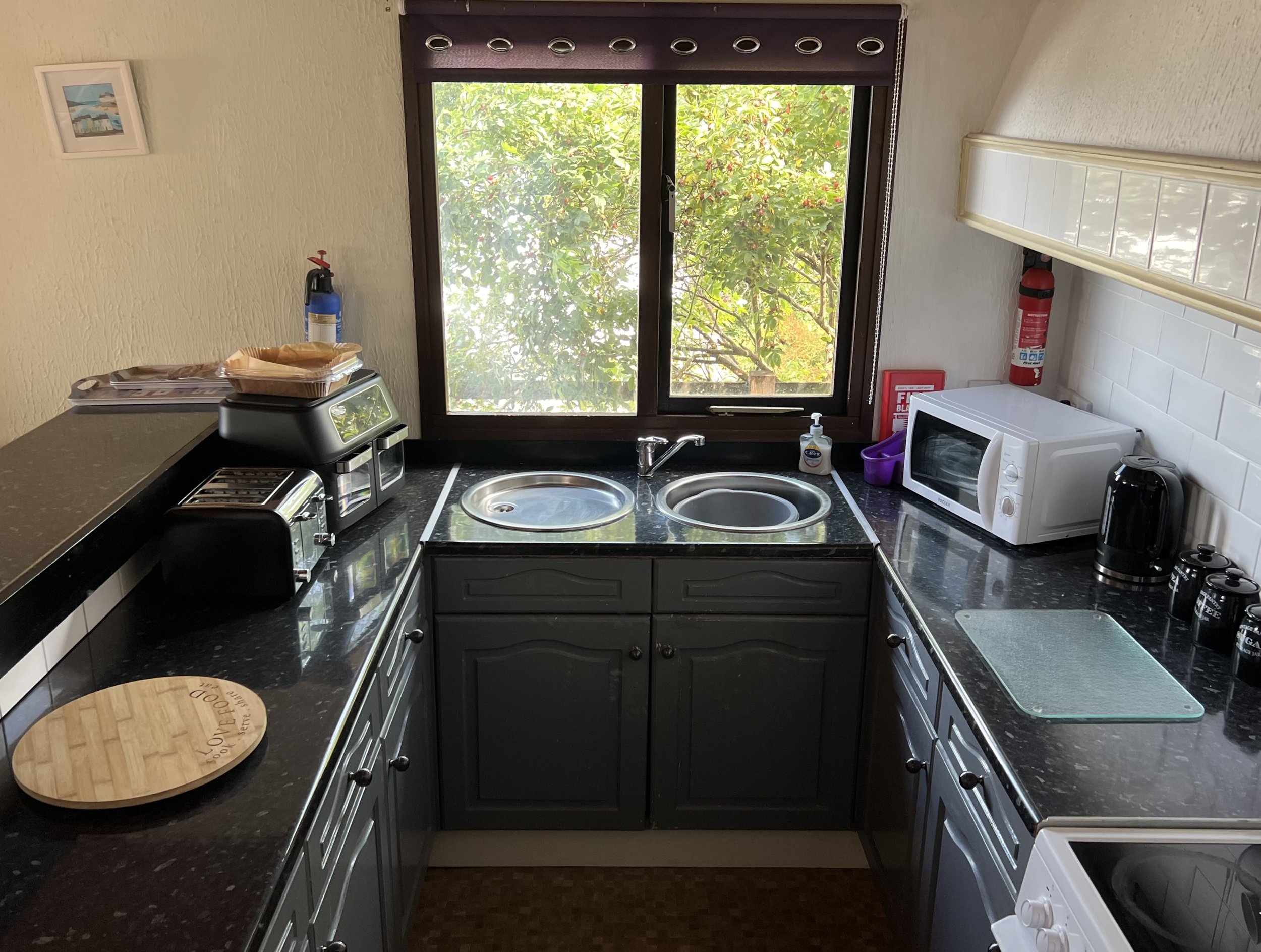 Kitchen with a window showing greenery outside, black countertops, and dark cabinets. There is a microwave, toaster, coffee maker, and fire extinguisher on the counter. A wooden cutting board is on the left side.