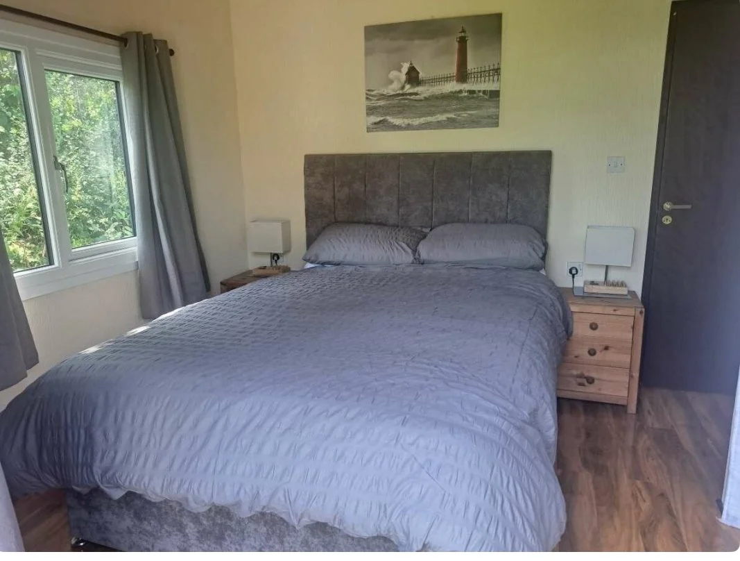 A bedroom featuring a bed with gray bedding and two matching pillows, wooden nightstands with lamps, a window with gray curtains, a lighthouse painting above the bed, and a wooden door on the right.