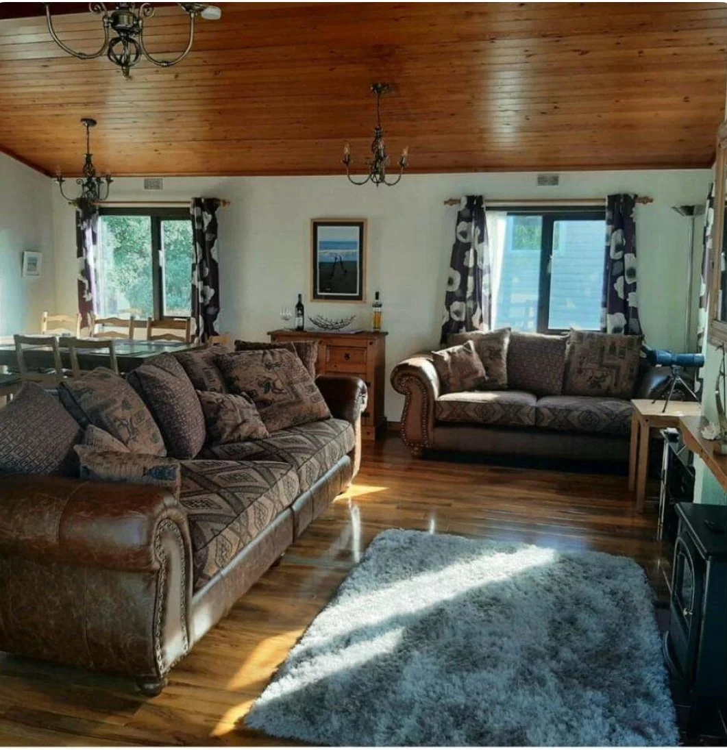Living room with wooden ceiling, two sofas with patterned cushions, a shaggy white rug, wooden furniture, windows with patterned curtains, and a telescope in the corner.