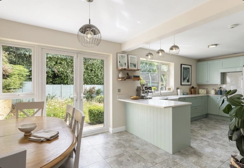 Bright kitchen with pale green cabinets, a white countertop island, and large windows and glass door overlooking a garden.