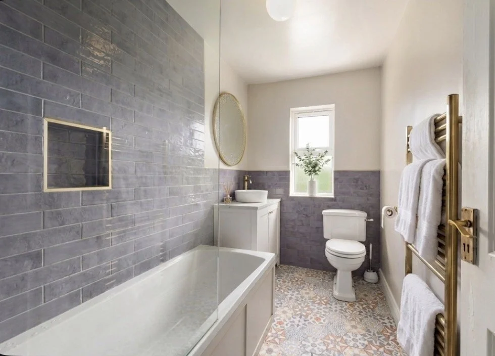 Modern bathroom with gray tiled wall, white bathtub, mirror, small sink cabinet, toilet, window with plant, and gold towel radiator with white towels.