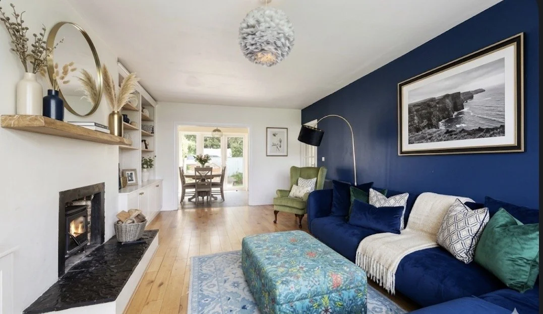 Living room with a blue accent wall, navy sofa, green armchair, and wooden flooring. Fireplace with black hearth, shelves with decorative items, a round mirror, and a light fixture overhead. Dining area with a table and chairs visible in the background.