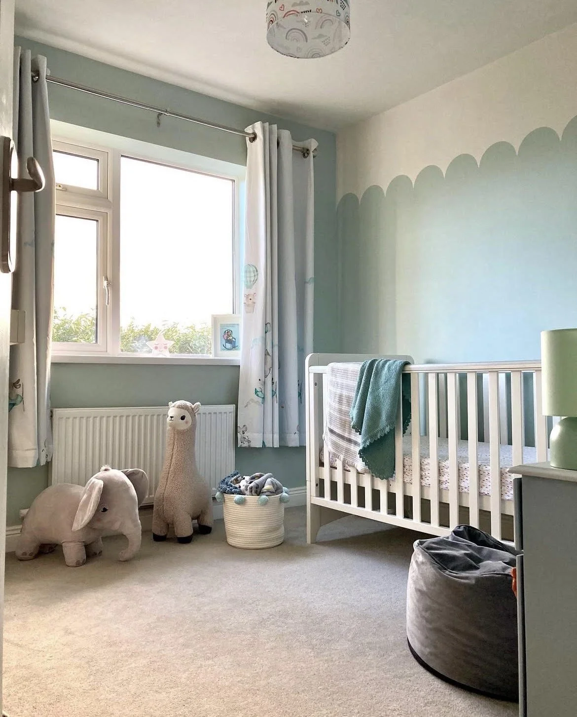 A cozy baby nursery with a white crib, a stuffed llama and a stuffed elephant on the beige carpet, a white basket filled with baby clothes, a green lamp on a small table, and children's drawings on the window sill. Light streams in through a large window with white curtains.