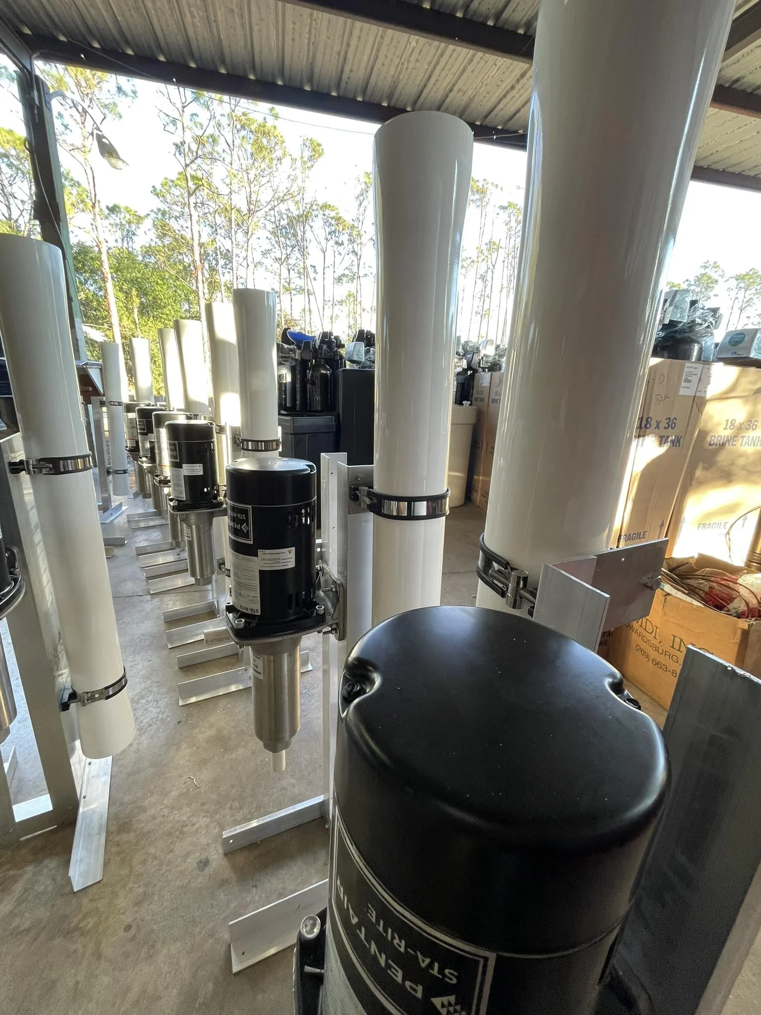 Shelves with water filtration components, including large white vertical filters, black motors, and other equipment, in a storage area with natural light and cardboard boxes in the background.