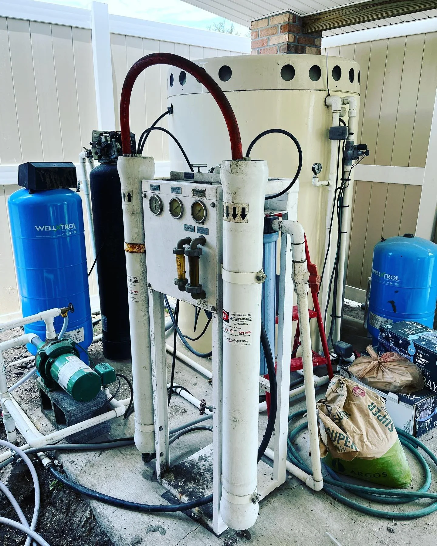 A backyard water filtration system with various pipes, gauges, and tanks. The system is installed on a concrete slab near a beige house wall with vinyl siding and a partial brick column.