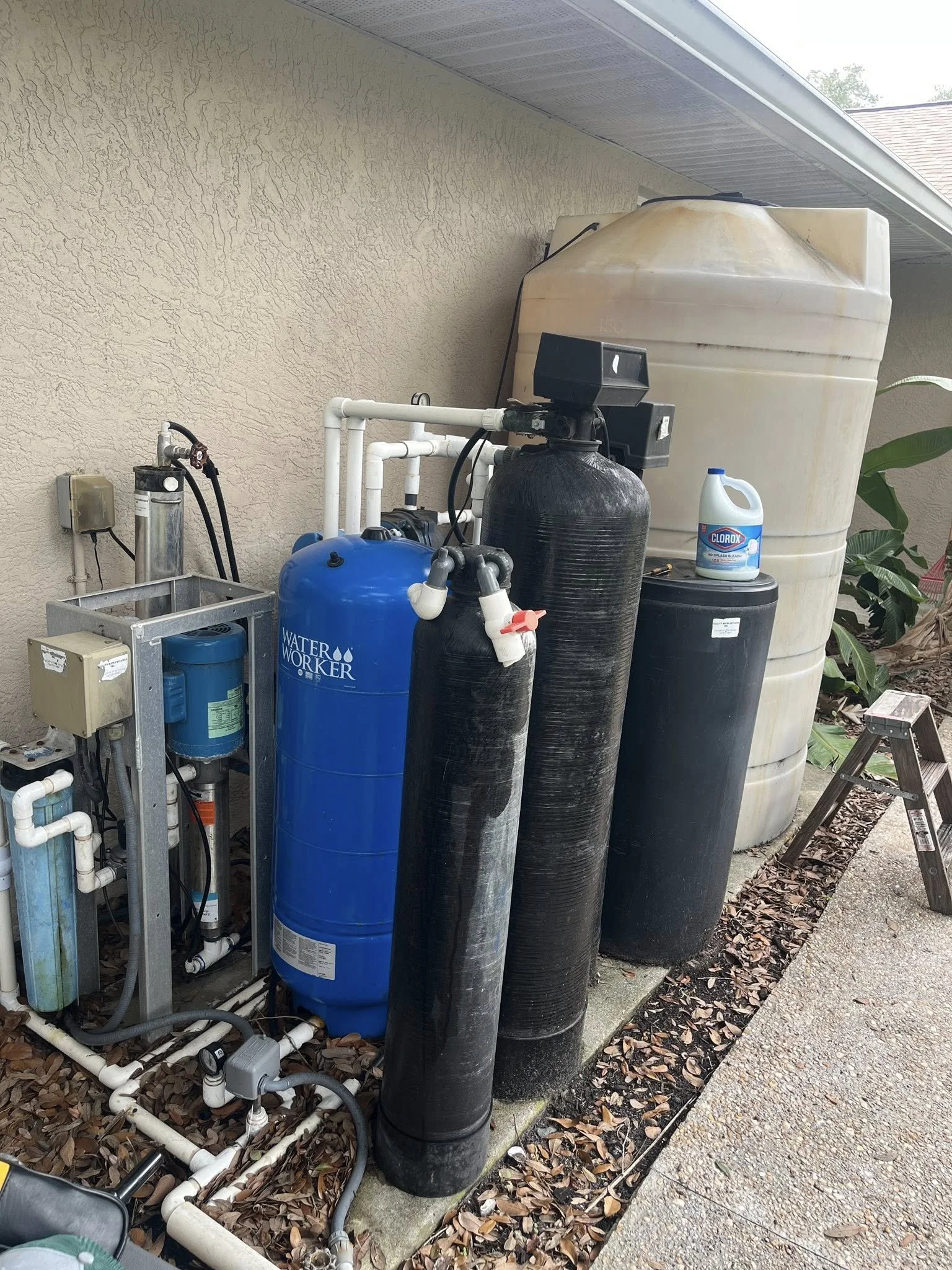 Outside view of a water filtration system with large black and blue tanks, white piping, a water softener, a large freshwater tank, a Clorox disinfectant bottle, and some greenery, located beside a house wall and walkway.