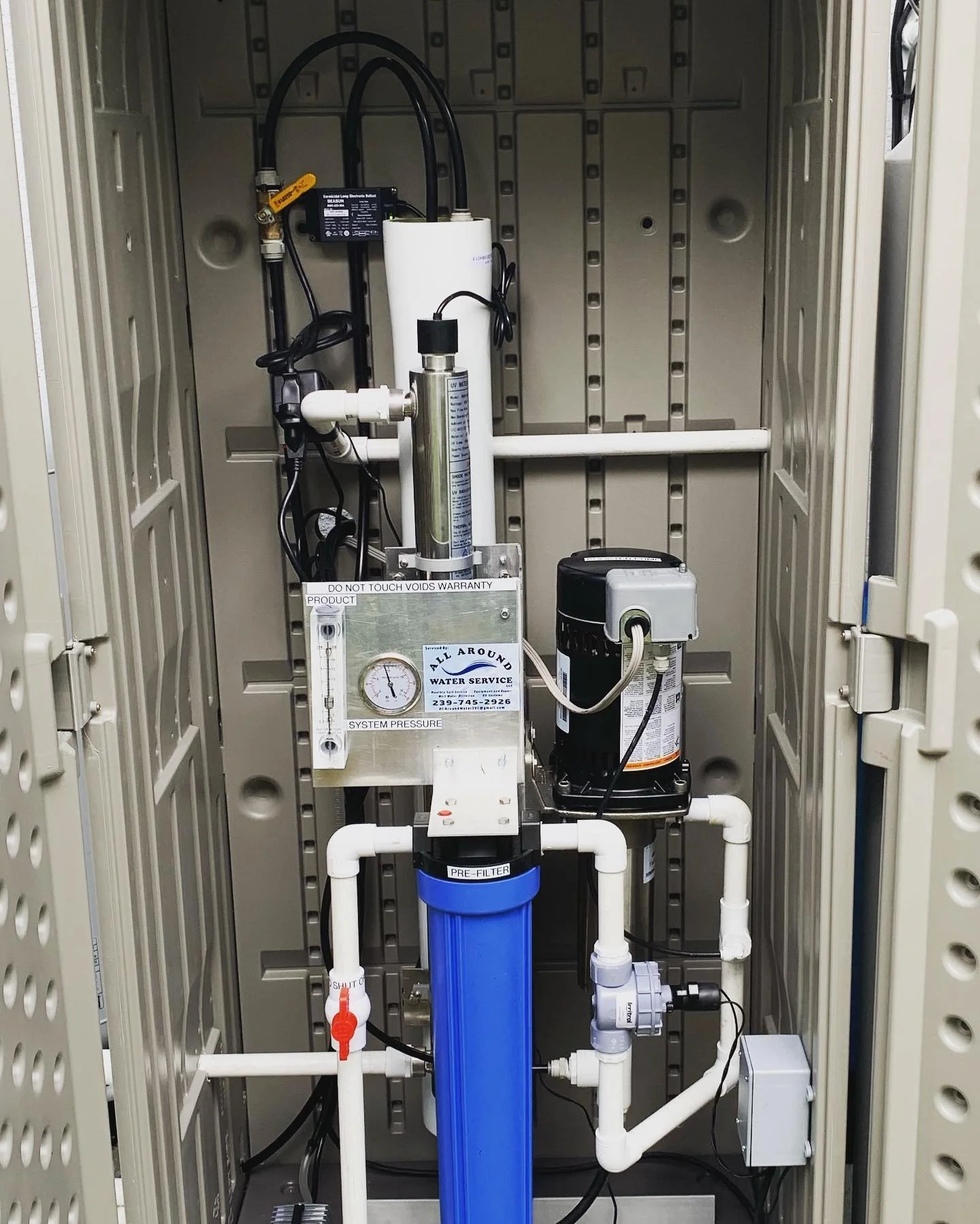 Inside an industrial water filter system with white and blue pipes, a pressure gauge, and black electrical components, enclosed in a beige metal cabinet.