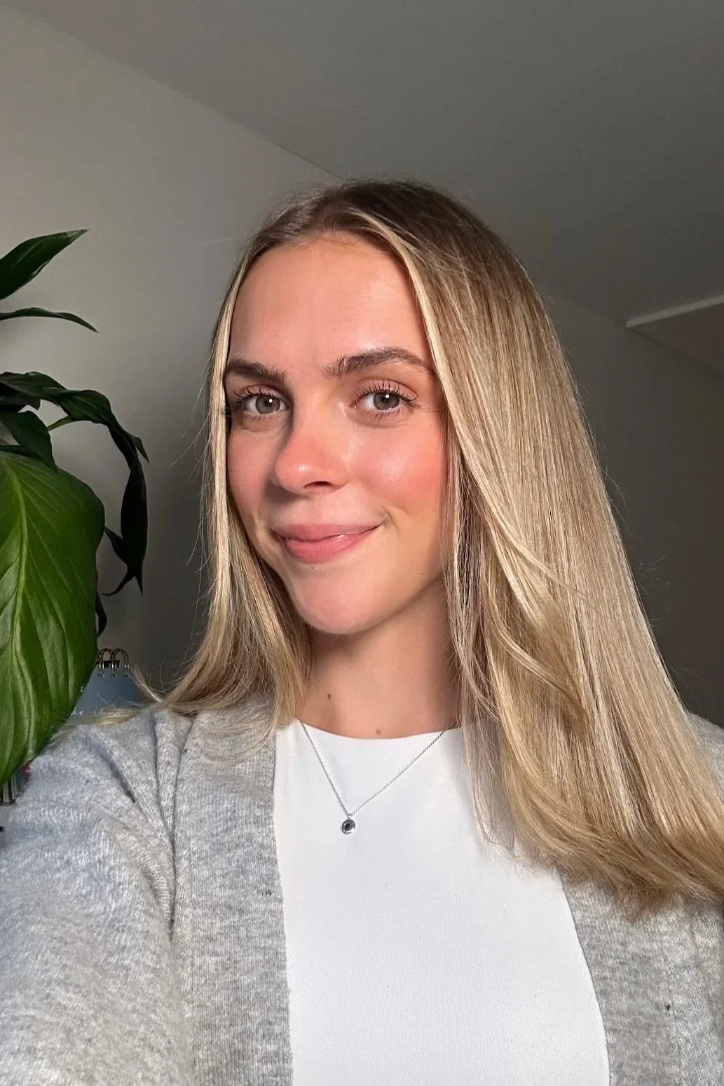 A young woman with long blonde hair, light makeup, and a smile taking a selfie indoors next to a green plant.
