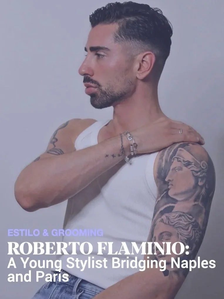 A man with styled dark hair and tattoos, wearing a white tank top and jewelry, looking to the left, with his right arm across his chest and hand on his shoulder.