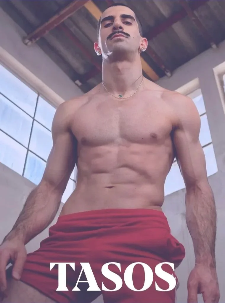 A shirtless young man with a mustache and earrings, wearing red shorts, looks downward with a serious expression inside a building with large windows and a wooden ceiling, with the word "TASOS" overlayed at the bottom.