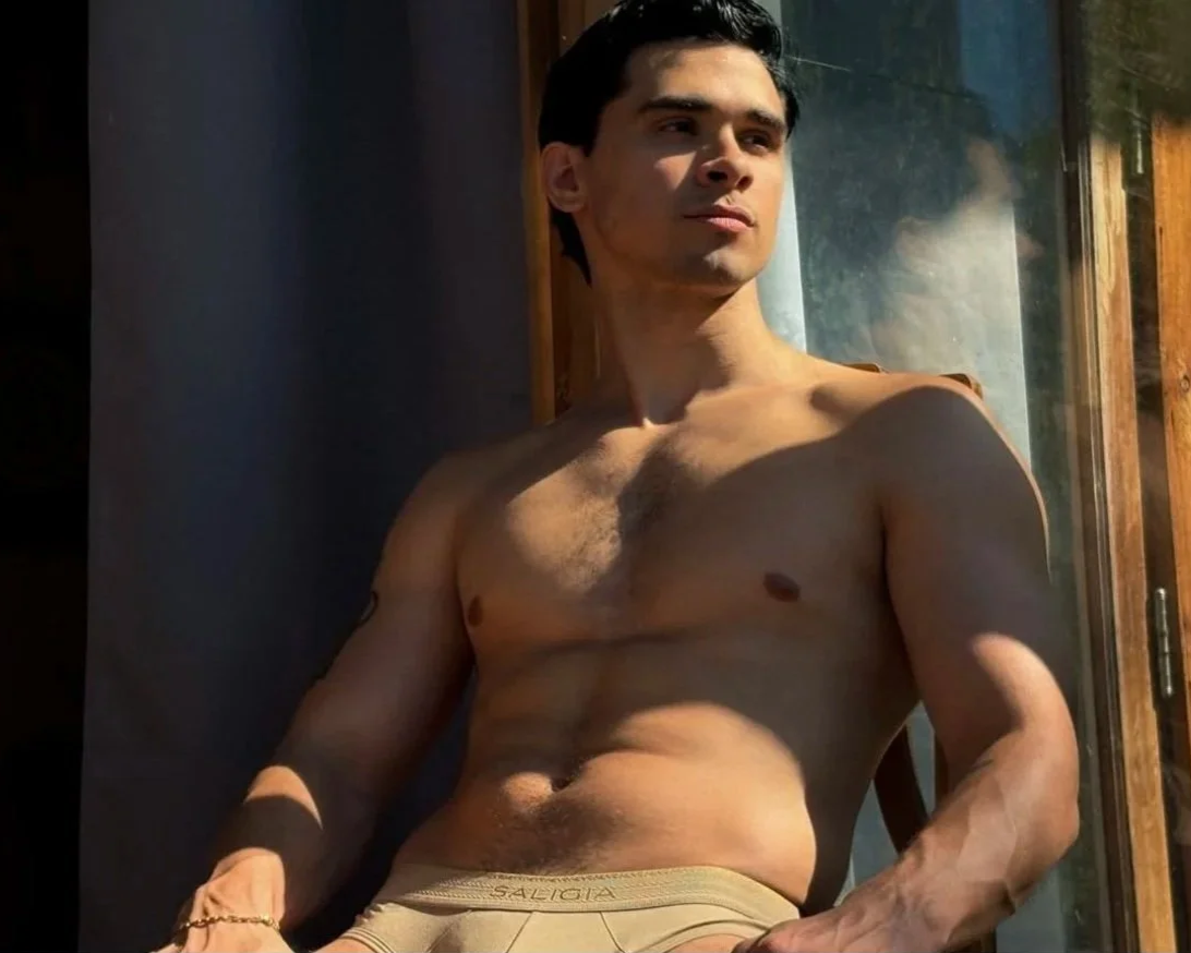 A shirtless young man with short dark hair wearing Saligia underwear and looking outside through a glass window, with sunlight illuminating his face and torso.