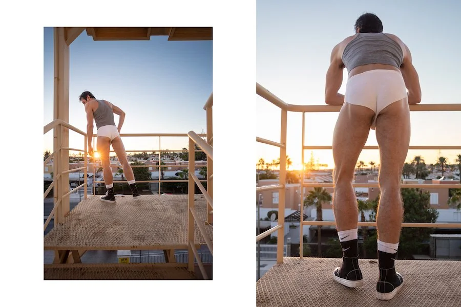 Fit man standing on a balcony, watching the sun set. He wears a white underwear that bares part of his glutes. Photo by @papagnimeca