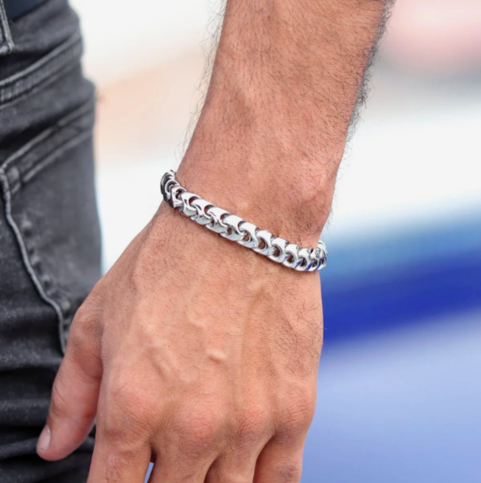 Close-up of a person's wrist wearing a silver chain bracelet, with part of their hand and arm visible, wearing a black shirt sleeve and black jeans.
