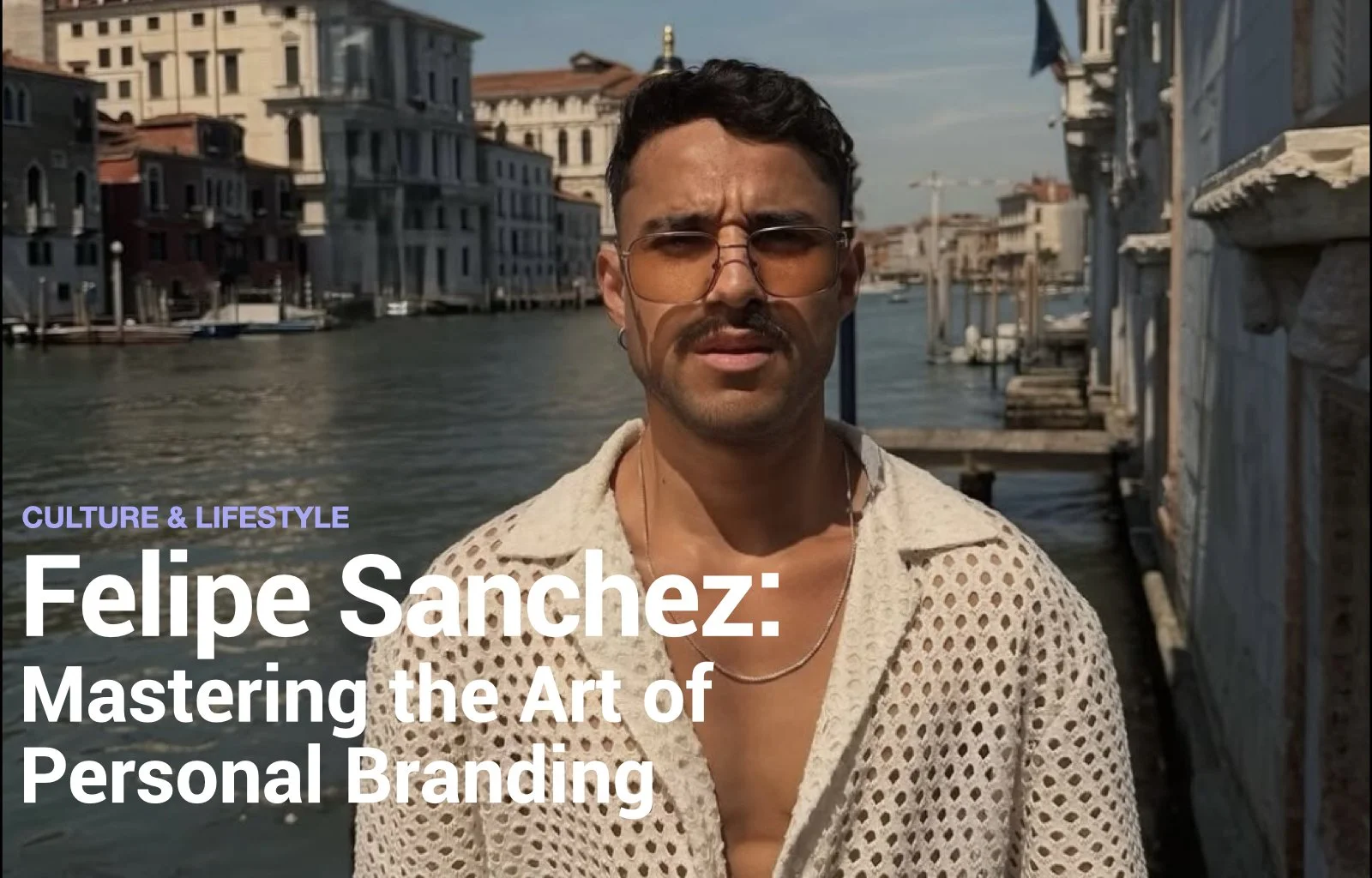A man with dark hair, a mustache, and brown-tinted glasses stands near a canal with buildings and boats in the background, during daytime.