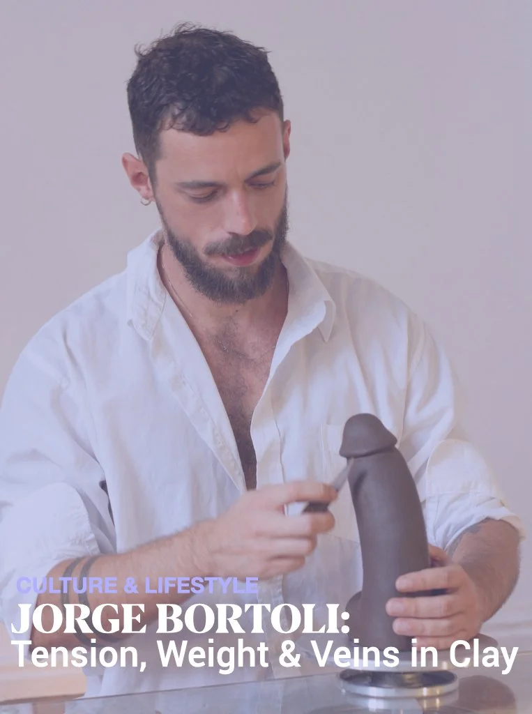 A man with dark hair, beard, and mustache wearing a white shirt, holding a clay sculpture of a penis with one hand and a sculpting tool with the other, in a minimalistic room.