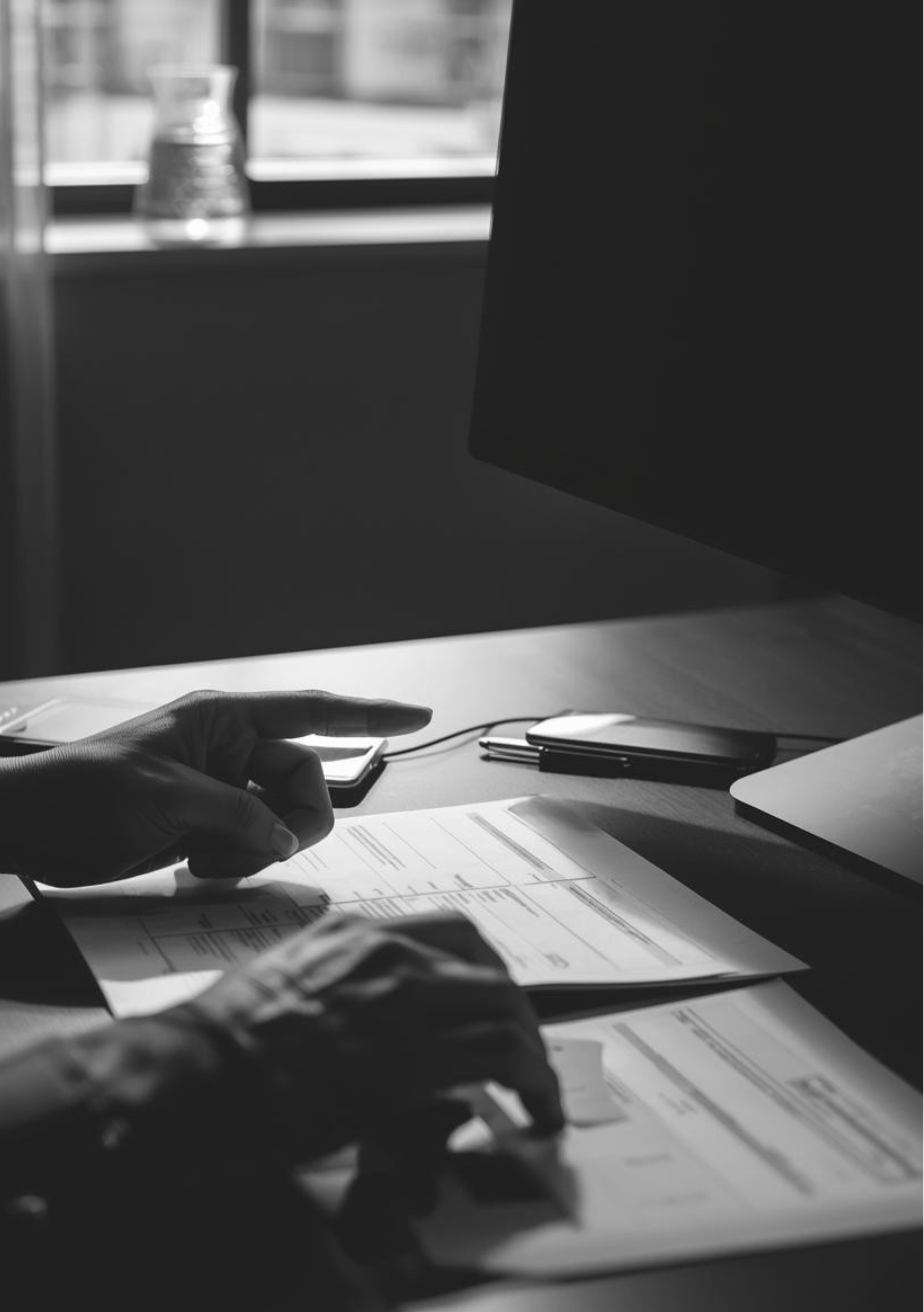 A person working at a desk during daylight, with documents, a computer monitor, a mouse, and a phone on the desk, and a window in the background.