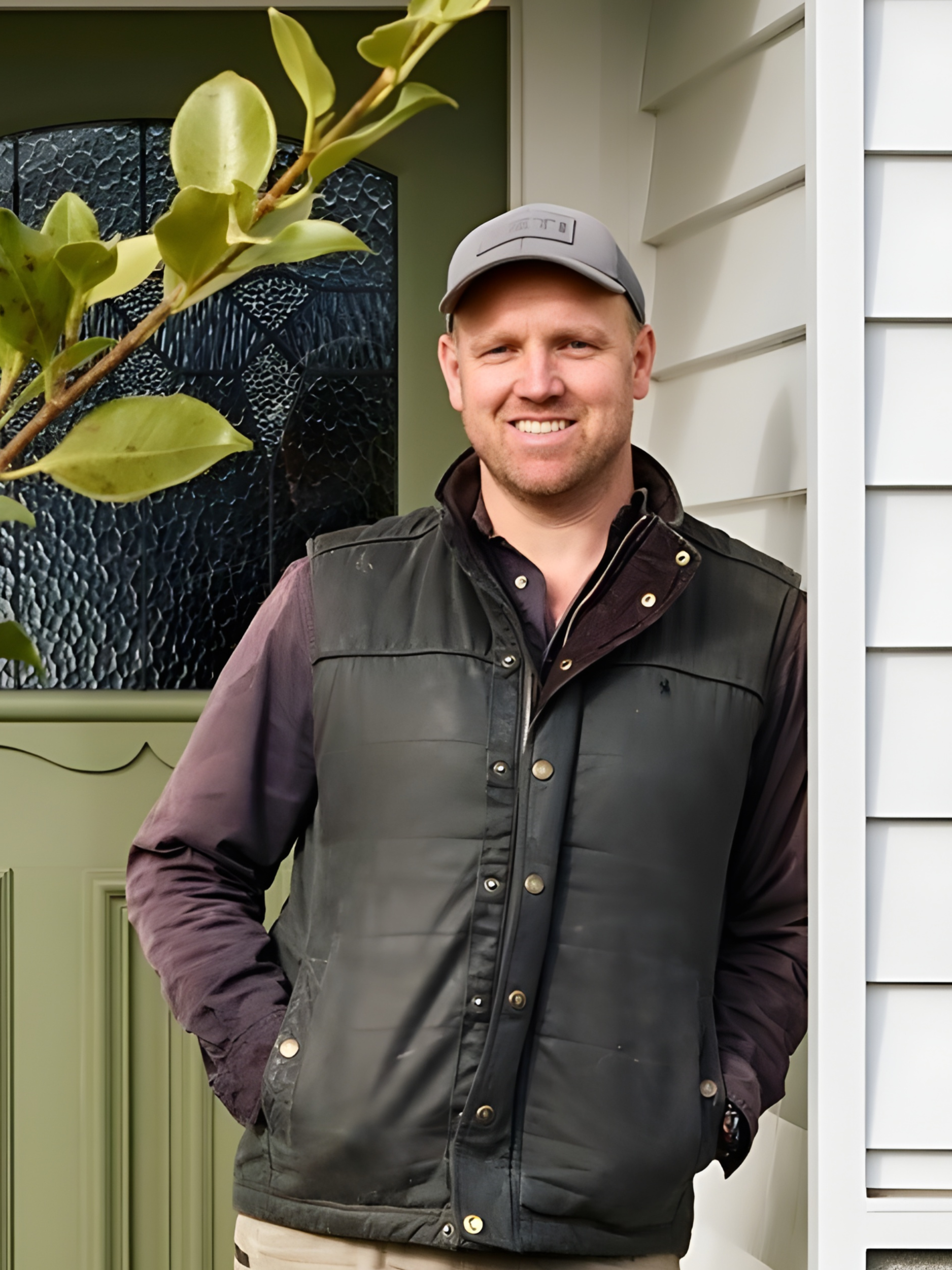 A man standing outdoors in front of a house, smiling, wearing a gray cap, dark vest, and a black watch.