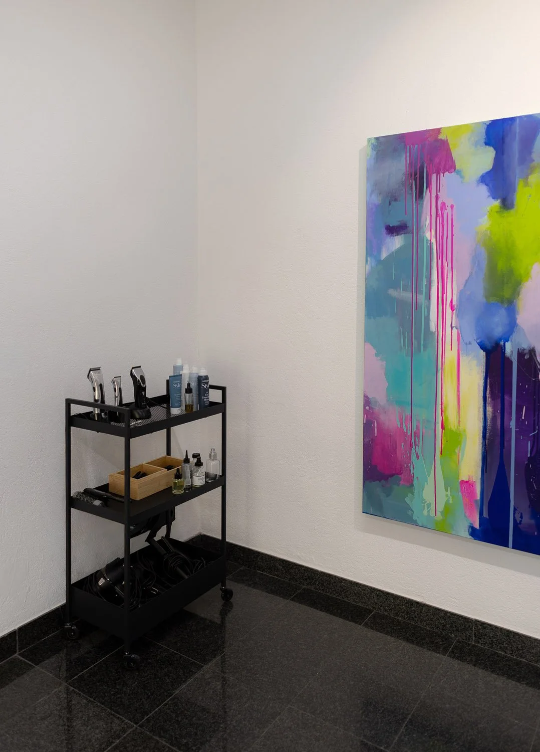 Eine Ecke in einem Kunststudio mit einem schwarzen Rollwagen, auf dem Haarentfernungsmittel, Rasierer und Pflegeprodukte stehen. Auf der Wand hängt ein großes, buntes abstraktes Gemälde.