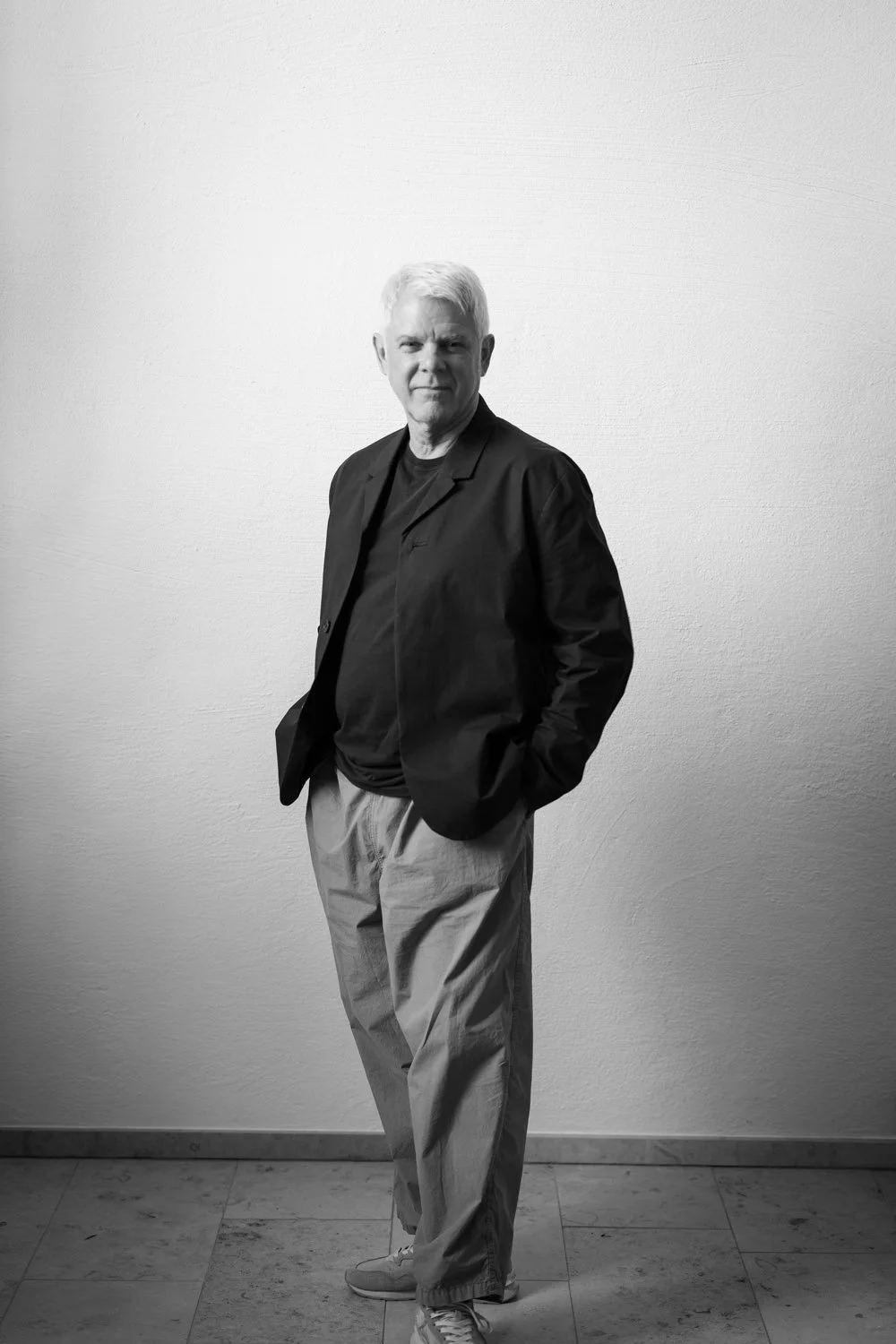 Schwarz-weiß-Foto eines älteren Mannes, der in lässiger Kleidung vor einer weißen Wand steht, mit den Händen in den Taschen.