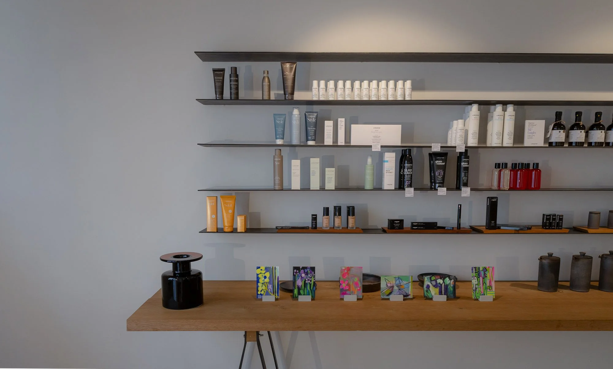 Regal mit Kosmetikprodukten, darunter Cremes, Shampoos und Flüssigkeiten, sowie gemalte Postkarten auf einem Holztisch.