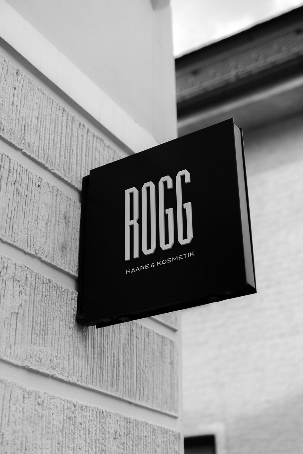 Schwarzes Schild mit weißen Buchstaben, das auf eine Kosmetik- und Friseursalons namens 'ROG' hinweist, an einer weißen Ziegelwand angebracht.