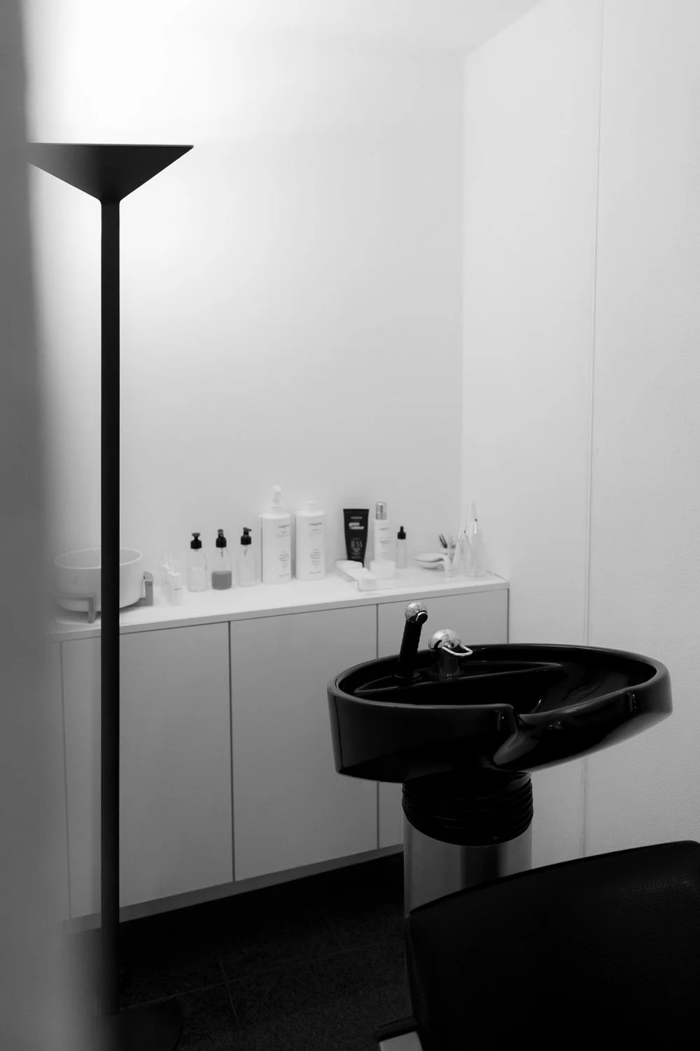 Schwarze Waschbecken im Behandlungsraum mit weißen Regalen und Medikamenten. Einschließlich eines Stehlamps, einer schwarzen Behandlungsliege und weißen Wandflächen.