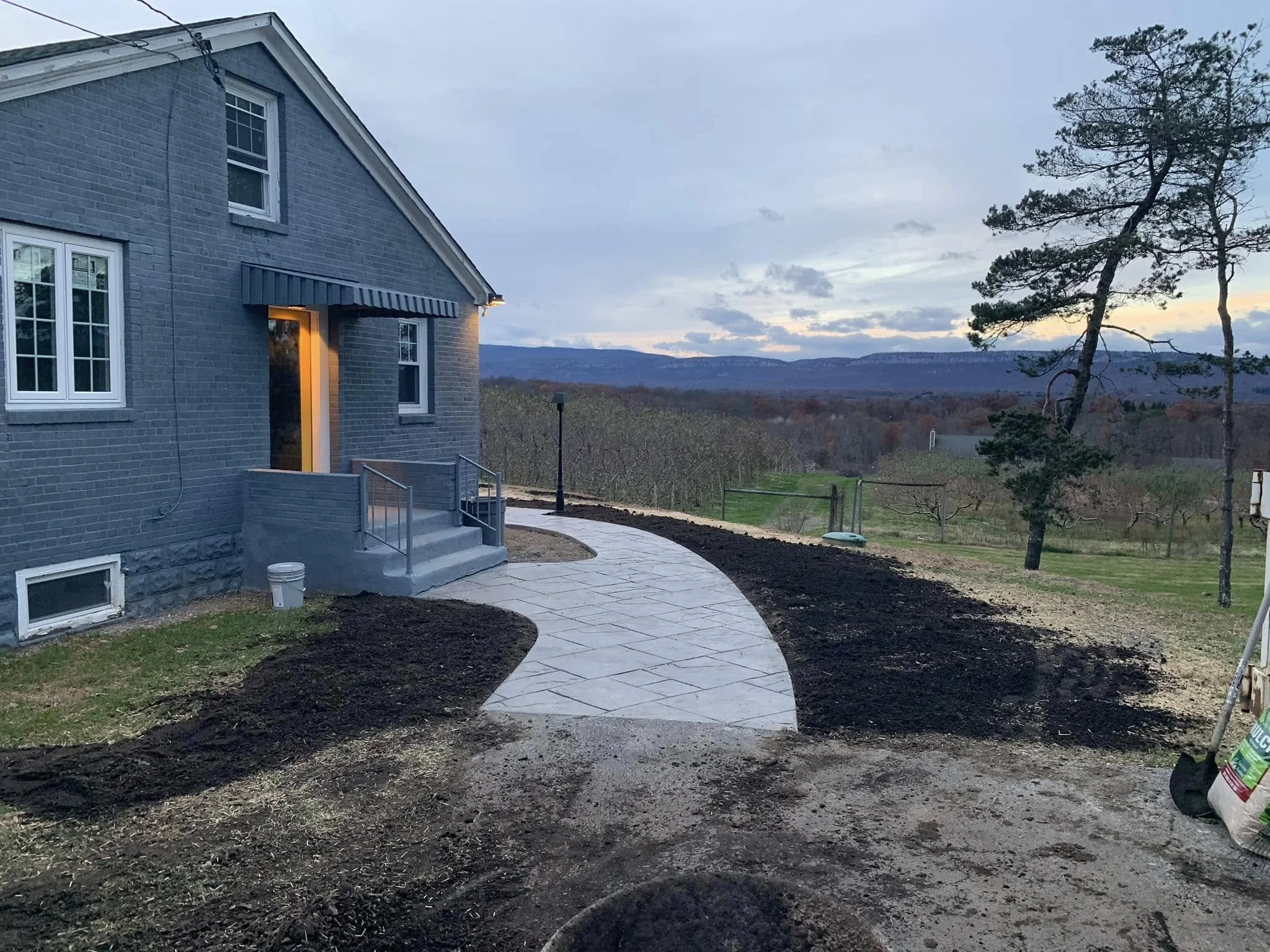 Backyard of renovated house in hudson valley with new paved walkway, a small set of stairs at the entrance, lawn, and trees with mountains in the distance, during dusk.