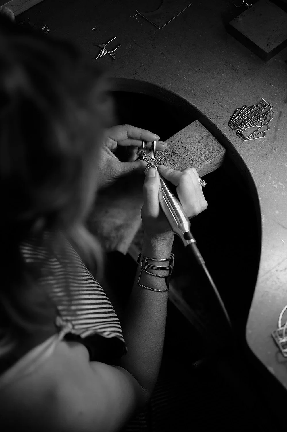A woman working on jewelry with a rotary tool at a jeweler's workstation, surrounded by metal clips and other small tools in a black-and-white photo.