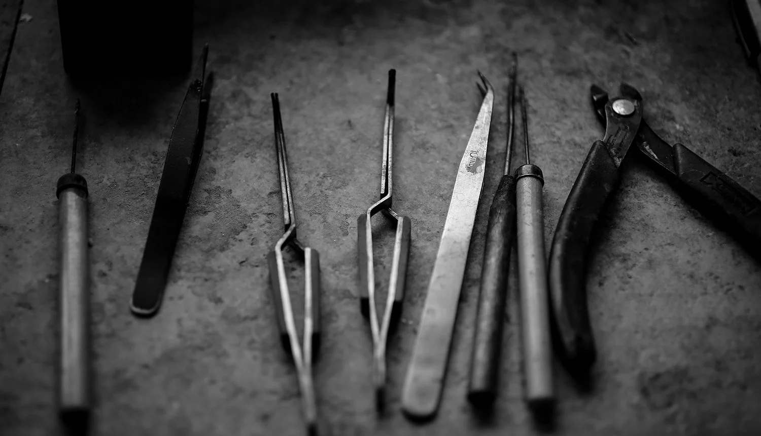 A collection of black and white hand tools arranged on a rough surface, including various types of pliers, tweezers, and a file.