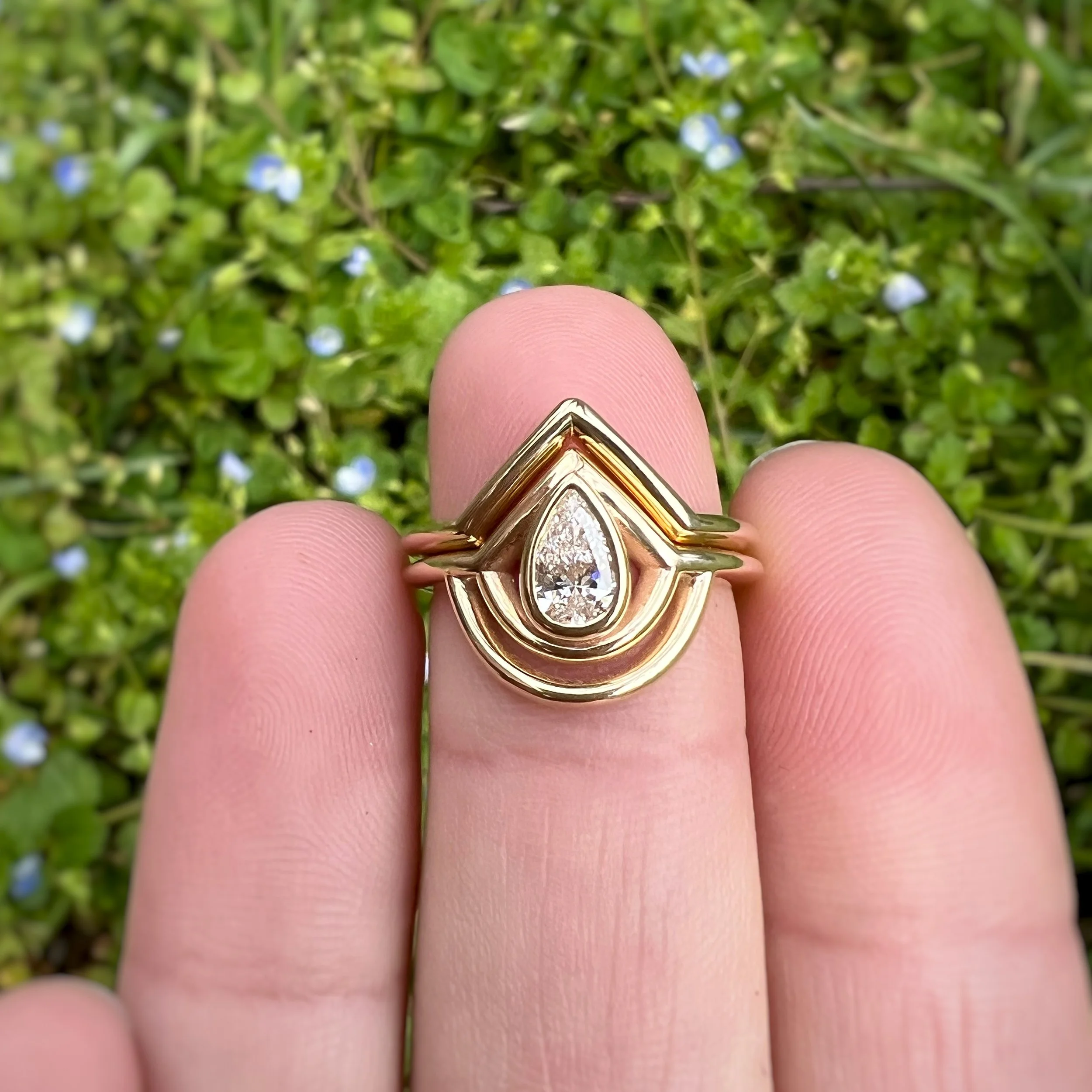 Close-up of a gold ring with a teardrop-shaped diamond, worn on a person's finger, with green foliage and small blue flowers in the background.
